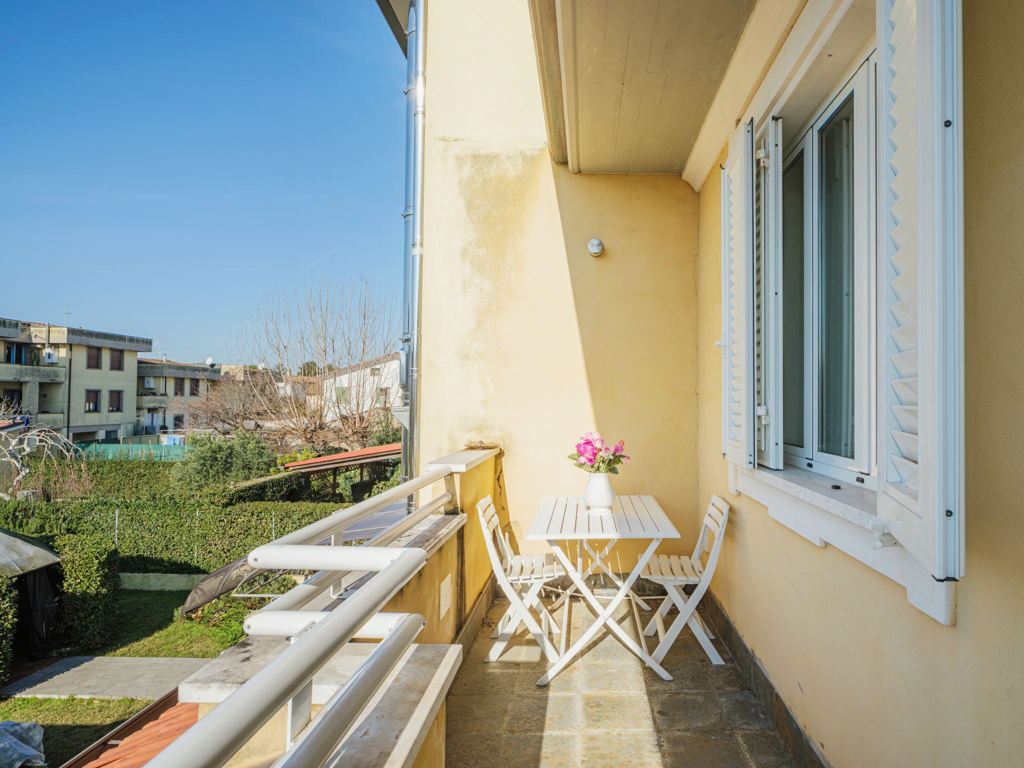 A balcony with a table and chairs, a small garden area, and a view of neighboring buildings.