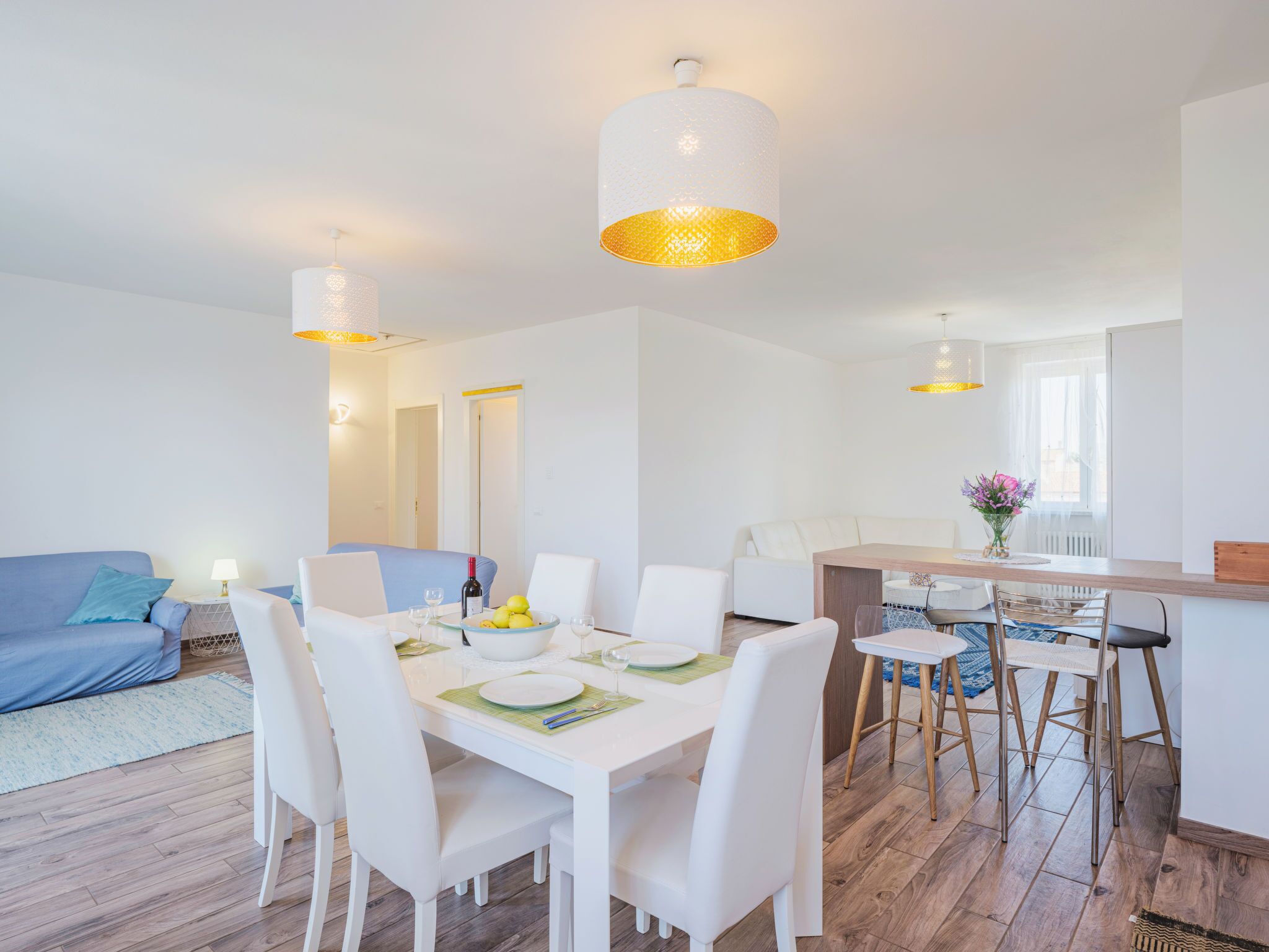 A modern living room with a dining area, featuring white furniture, a wooden dining table, and pendant lighting.