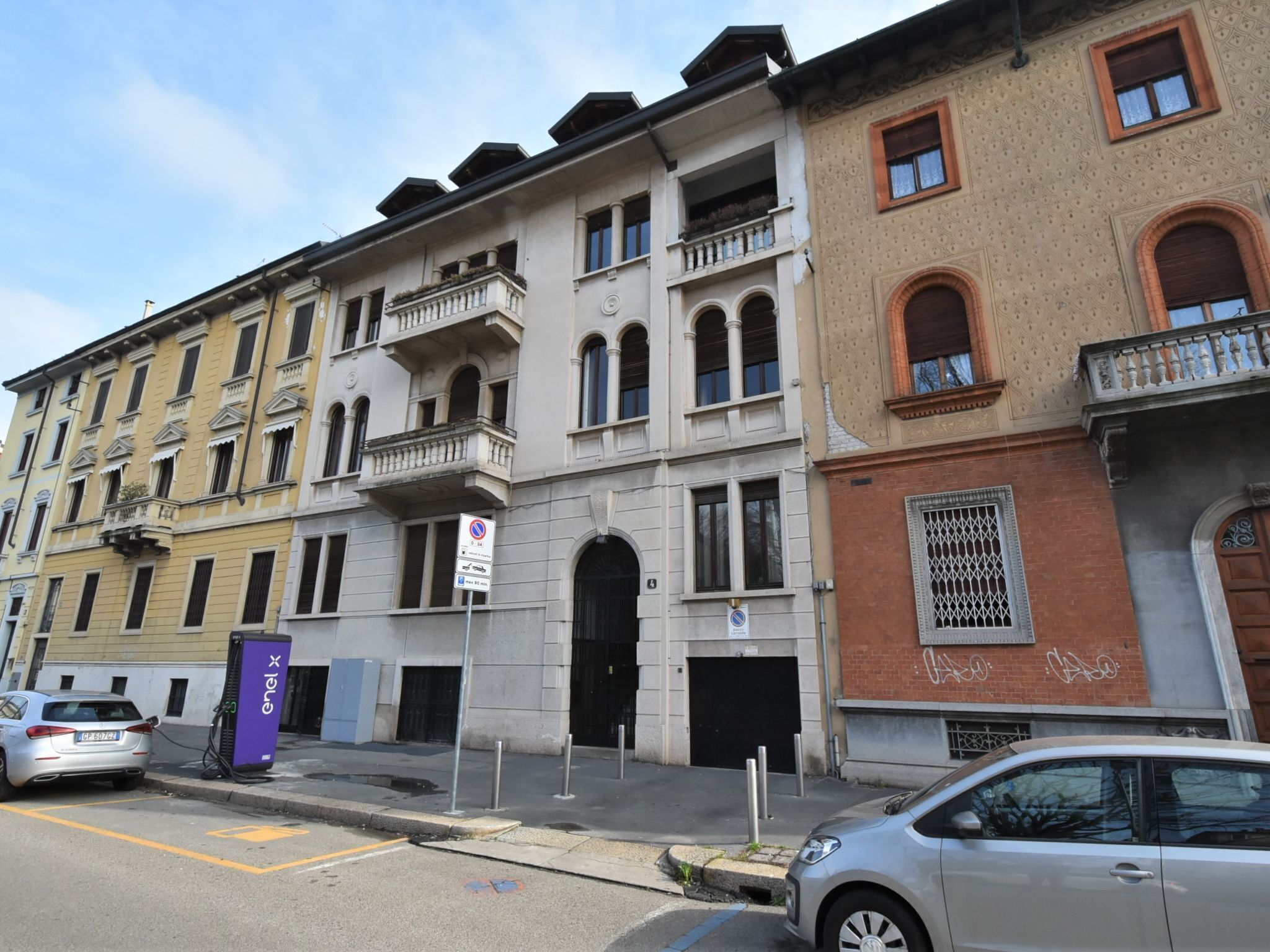 Una hilera de edificios de varios pisos con balcones, una calle con coches aparcados y un cielo despejado.