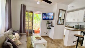 Ein modernes Wohnzimmer mit Sofa, Couchtisch und Deckenventilator. Ein Küchenbereich mit weißen Schränken und einer Mikrowelle ist sichtbar.