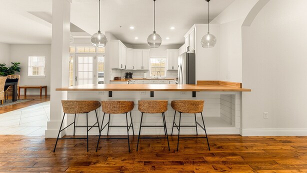 Uma cozinha moderna com armários brancos, uma ilha de madeira com banquetas, luminárias pendentes e uma janela grande.