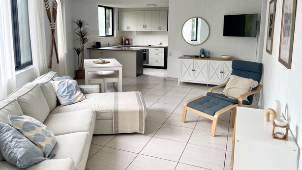 Ein modernes Wohnzimmer mit Sofa, Sessel, Esstisch und einer Küche im Hintergrund.