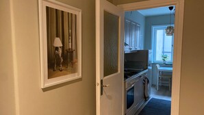 Un couloir mène à une cuisine comprenant un coin repas, une fenêtre avec une plante et une porte ornée d’un tableau encadré.