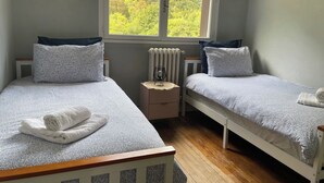 Ein Schlafzimmer mit zwei Betten, einem Fenster mit Blick auf Grünflächen, einem Heizkörper und einem Nachttisch mit Lampe.
