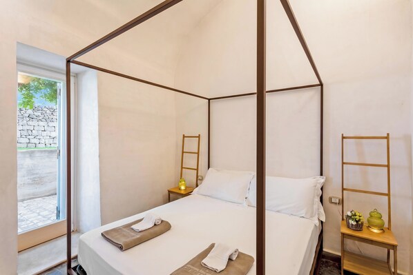 Un lit à baldaquin avec un matelas et des oreillers blancs, une table de chevet avec une lampe, et une petite étagère avec des objets décoratifs.