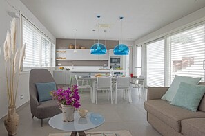 A modern living room with a sofa, armchair, and coffee table. There is a dining area with a table and chairs. The kitchen is visible in the background with white cabinets and a microwave.
