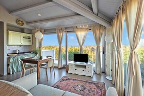 A living room with a sofa, a TV on a white cabinet, a dining table with chairs, and a kitchen area with a stove.