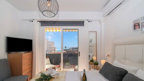A hotel room with a bed, a TV, a balcony with a view of buildings, and a dining area with a table set for two.