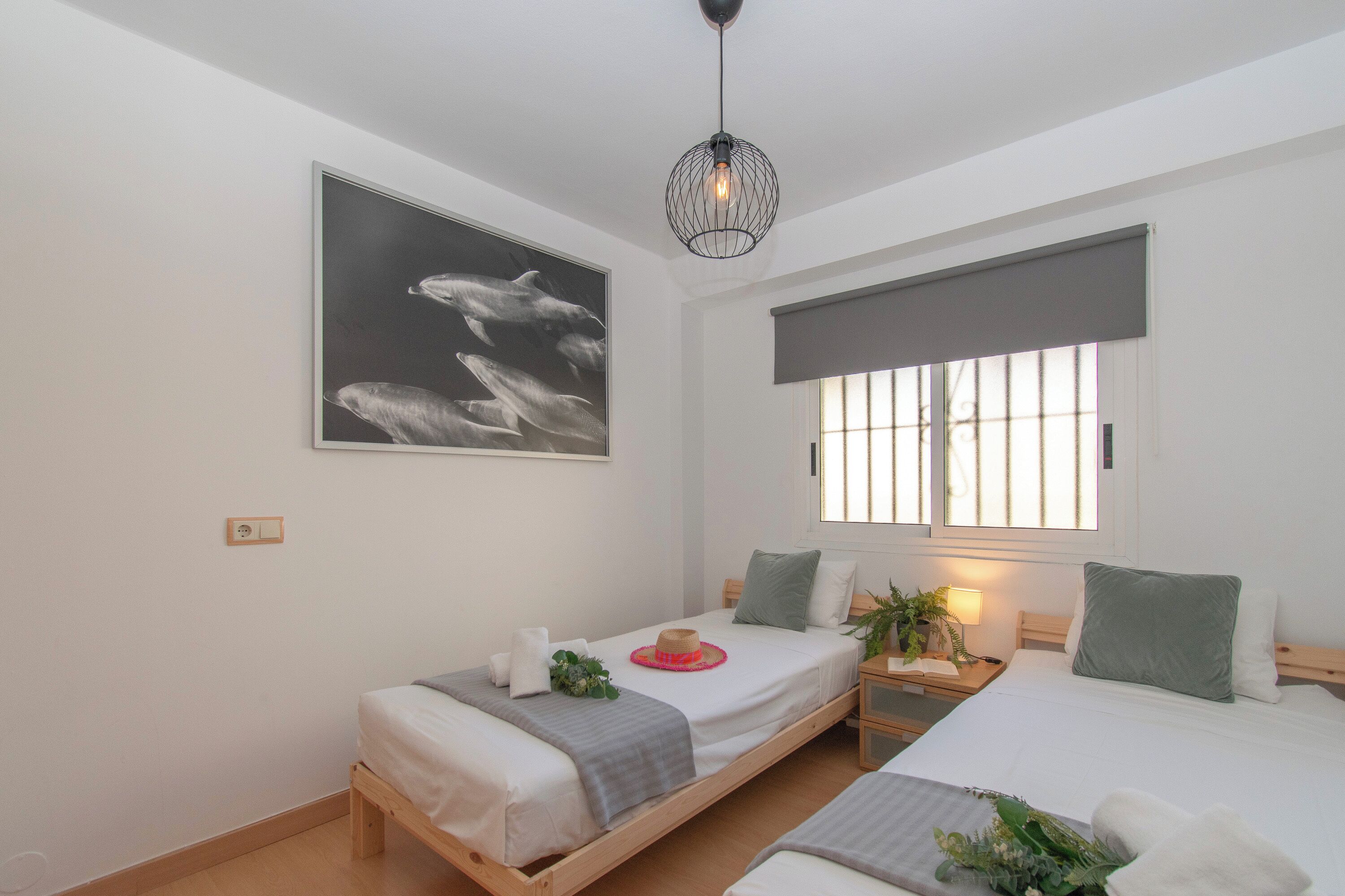 A bedroom with two beds, a window with blinds, a hanging light fixture, and a wall-mounted picture.