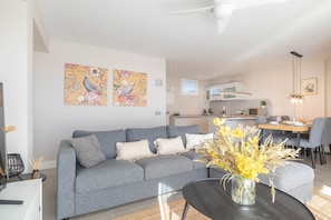 A modern living room with a grey sofa, a dining area with a table and chairs, and a kitchen in the background.