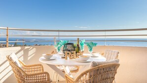 Terraza con sillas de mimbre, una mesa con platos, vasos y una botella, con vistas a la costa.