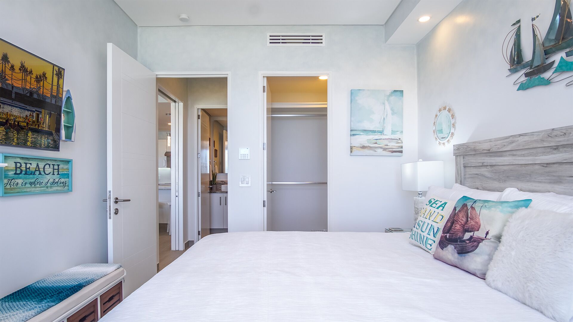 Une chambre à coucher comprenant un lit, un banc, une porte donnant sur une salle de bain et un mur orné d’un tableau et d’une affiche.