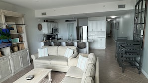 Un soggiorno con divano, tavolino e TV. C'è una cucina con frigorifero, microonde e pensili. Un cartello recita "BEACH HOUSE".