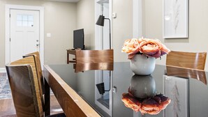 Un coin repas avec une table en verre, des chaises en bois, un vase avec des fleurs, une lampe noire et une porte en arrière-plan.