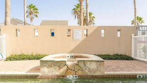 Un espace piscine agrémenté d’une fontaine, de palmiers et d’un mur de couleur beige percé de petites fenêtres.