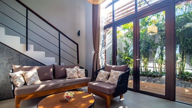 Ein modernes Wohnzimmer mit Sofa, Sessel und Couchtisch. Eine Treppe und große Fenster mit Blick ins Grüne sind zu sehen.