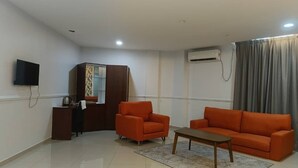 Un salon comprenant un canapé, un fauteuil et une petite table. On y trouve également un téléviseur fixé au mur, un meuble et un appareil de climatisation.