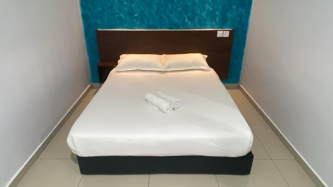 A neatly made bed with white linens and a rolled towel, a dark headboard, and a blue textured wall behind it.