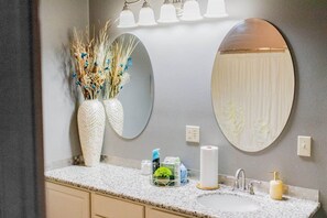 A bathroom with a granite countertop, two white vases with dried plants, a mirror with a circular frame, a sink with a gold faucet, and a soap dispenser.