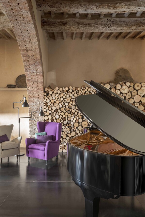A grand piano in a room with exposed brick walls, a stack of logs, and a modern chair. - Monteverdi Tuscany (Sarteano)