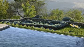 Relax in a serene infinity pool surrounded by a beautifully maintained garden and a stone wall, offering breathtaking views of a distant valley and water.