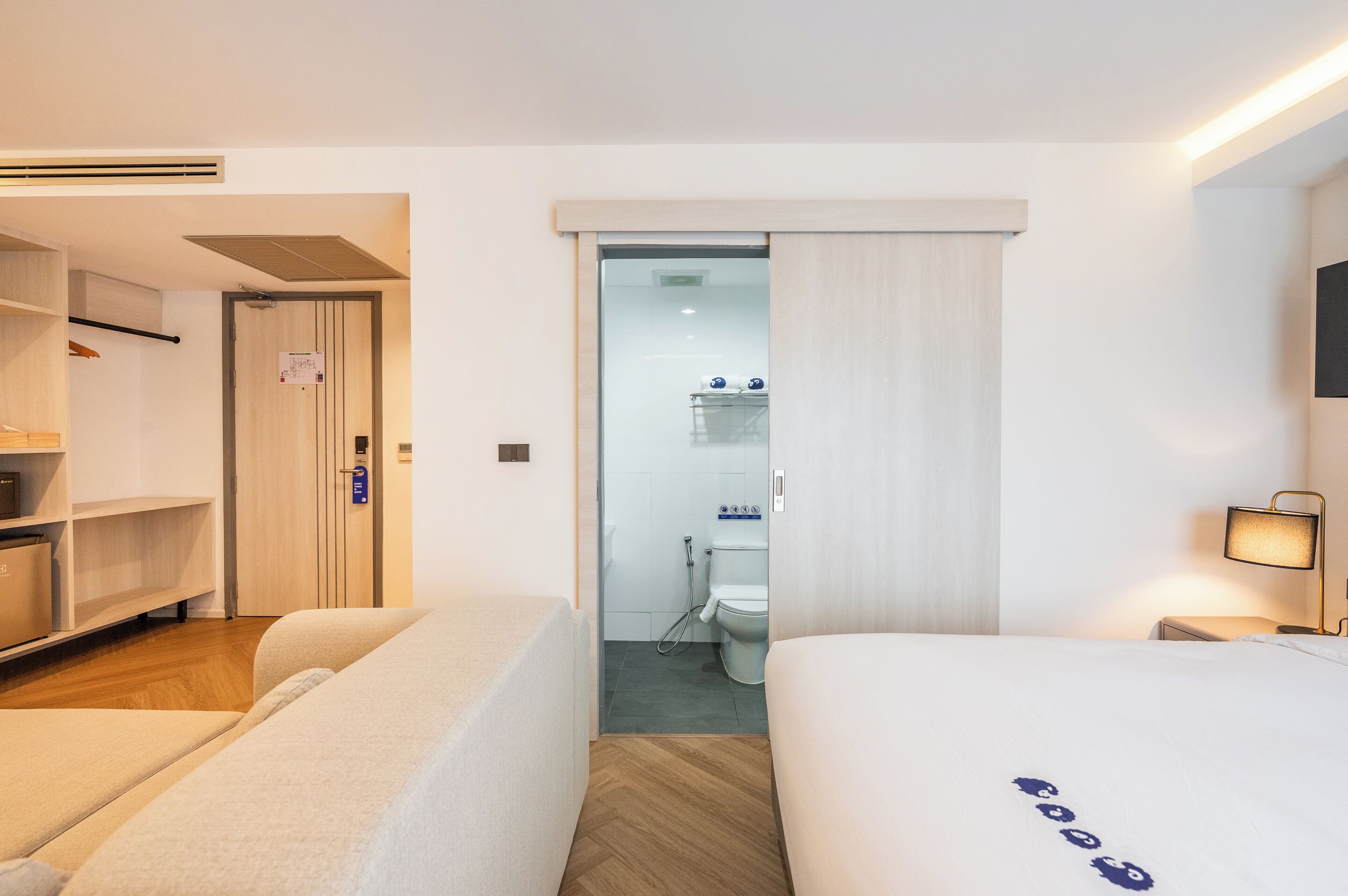 A hotel room with a bed, a bedside table with a lamp, a wardrobe, and a bathroom visible through a door.