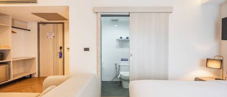 A hotel room with a bed, a bedside table with a lamp, a wardrobe, and a bathroom visible through a door.
