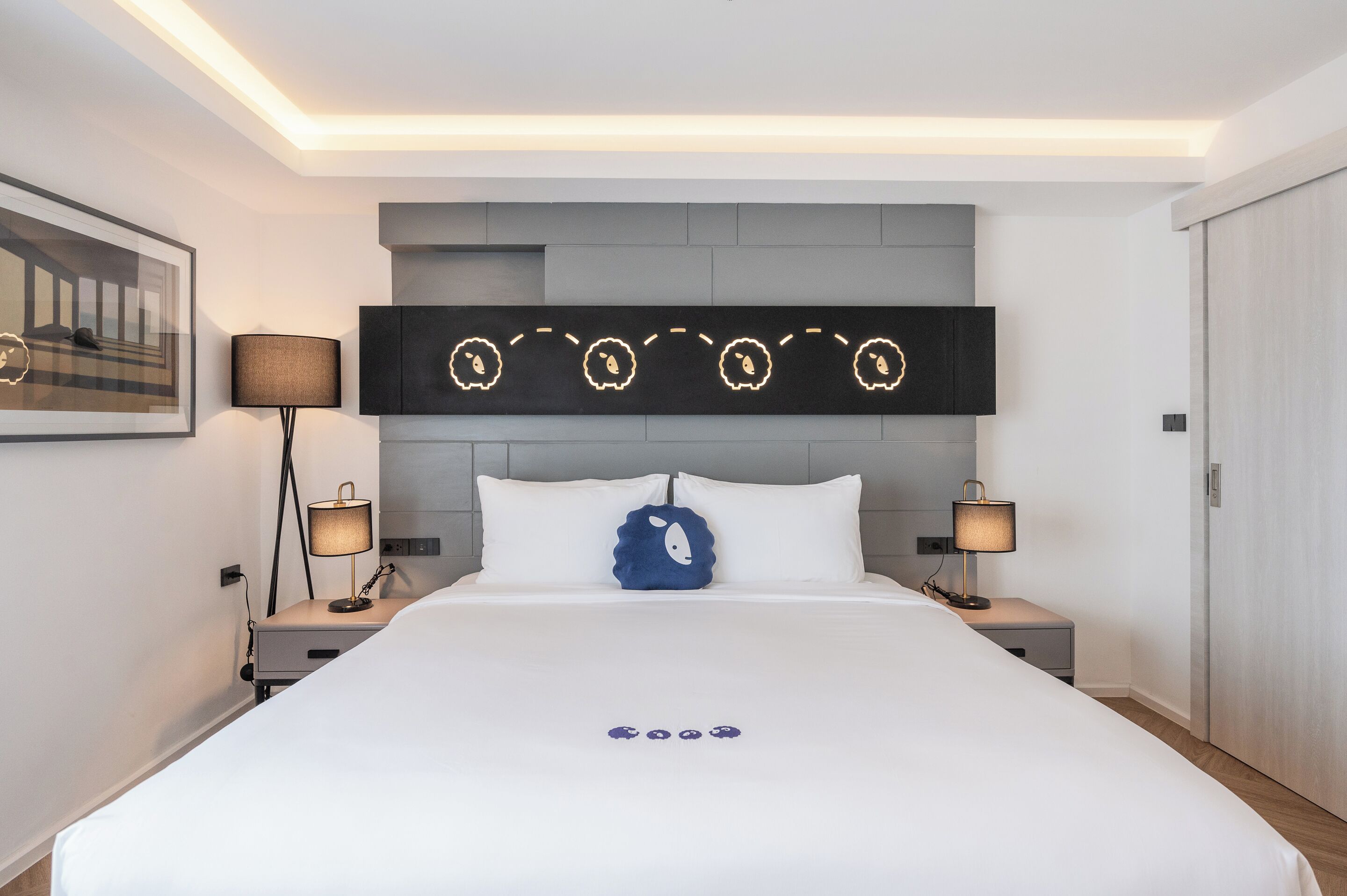 A modern hotel room with a large bed, two bedside tables, a headboard with decorative lights, and a framed picture on the wall.