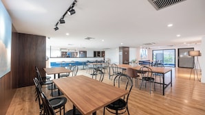 A spacious dining area with wooden tables and black chairs, a kitchen in the background, and a large window providing natural light.