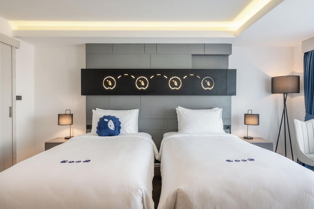 A hotel room with two beds, a modern headboard, bedside lamps, and a wall-mounted light fixture.