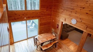 Interior de uma cabana de madeira com uma mesa de jantar, bancos, uma lareira e uma janela grande com vista para um deck.