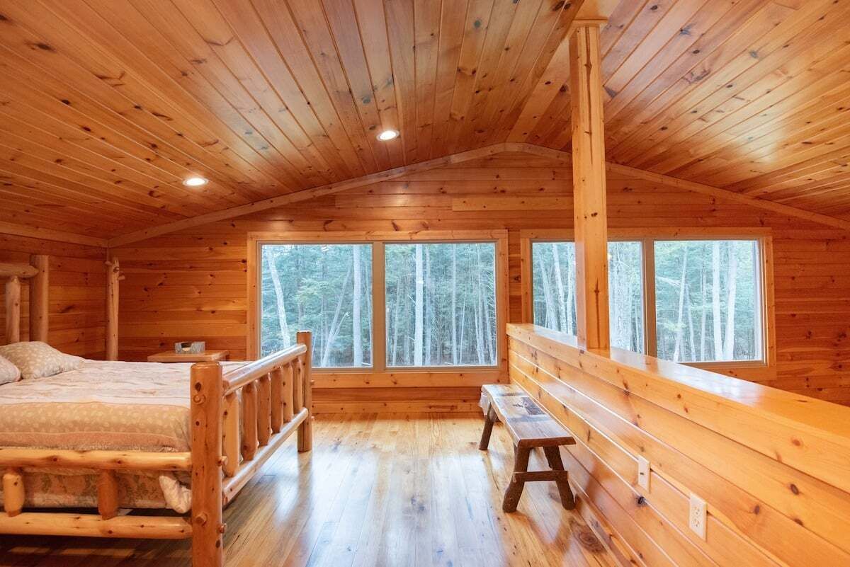 A log cabin bedroom with a wooden ceiling, a bed, a bench, and large windows.