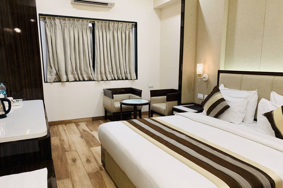 A hotel room with a large bed, a small table, a chair, and a desk with a kettle.