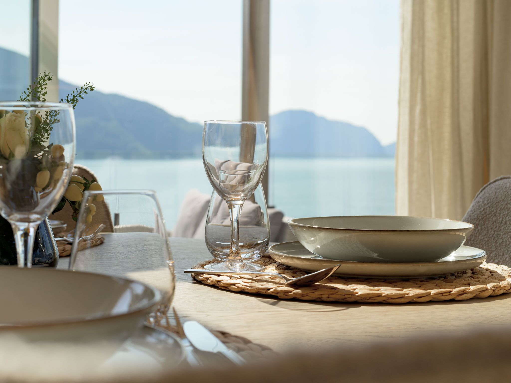 Una mesa de comedor con vista a un lago y montañas, dispuesta con platos, vasos y un bol.