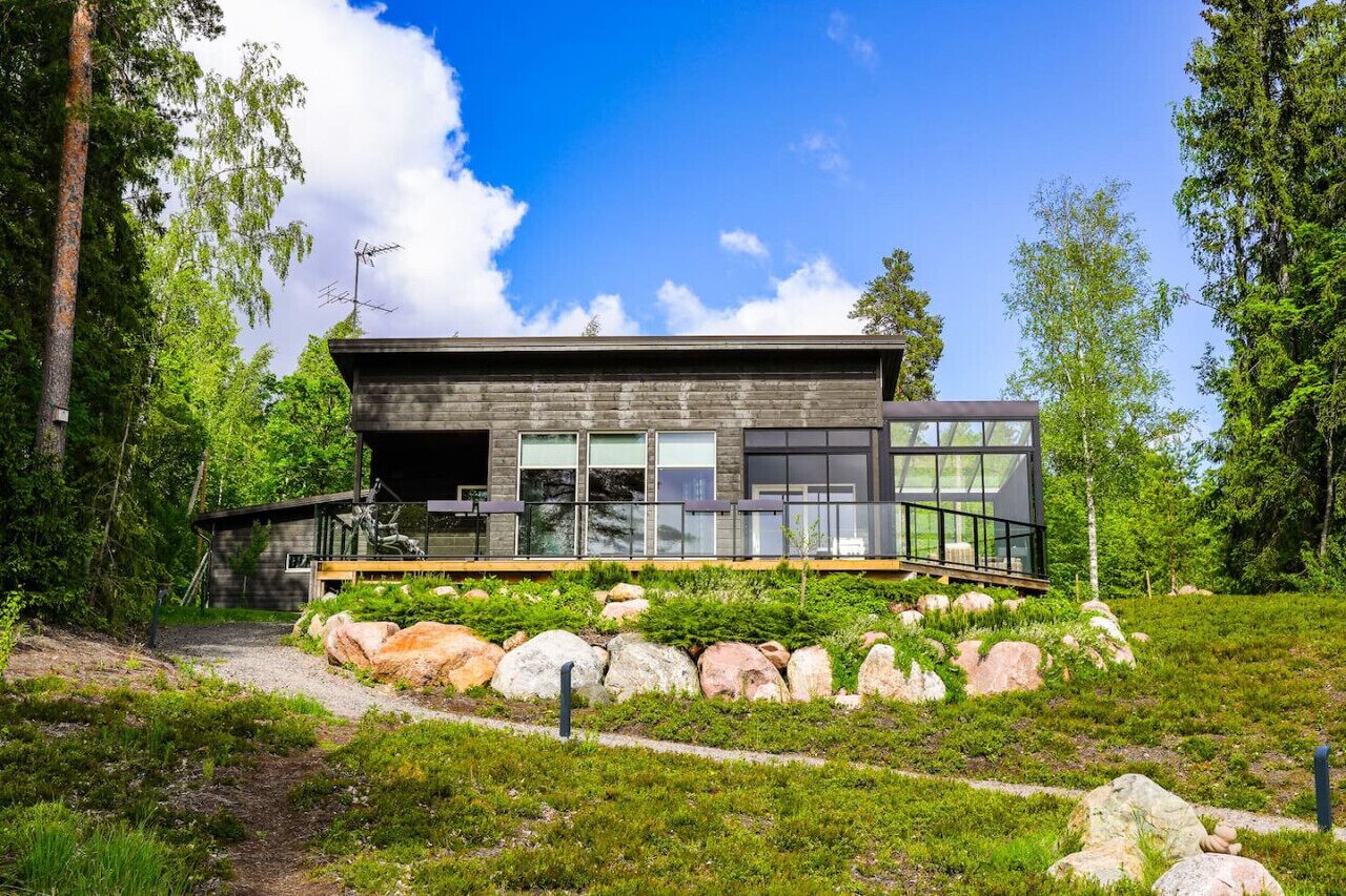 Una casa moderna de dos pisos con ventanas amplias, rodeada por un paisaje natural con rocas y árboles.