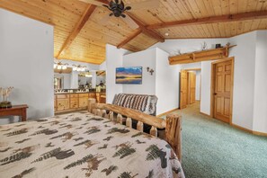Een slaapkamer met een groot bed, houten hoofdbord, houten plafond, een schilderij, een spiegel en een gang die naar een deur leidt.