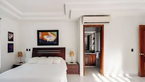 Hotelkamer met een groot bed, een houten hoofdbord, een schilderij boven het bed, twee nachtkastjes met lampen en een uitzicht op de keuken door een geopende deur.