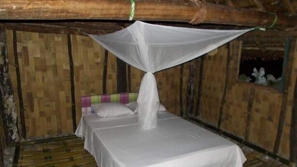 Un lit simple, protégé par une moustiquaire, dans une chambre rustique aux murs en bois et au plafond en bambou.