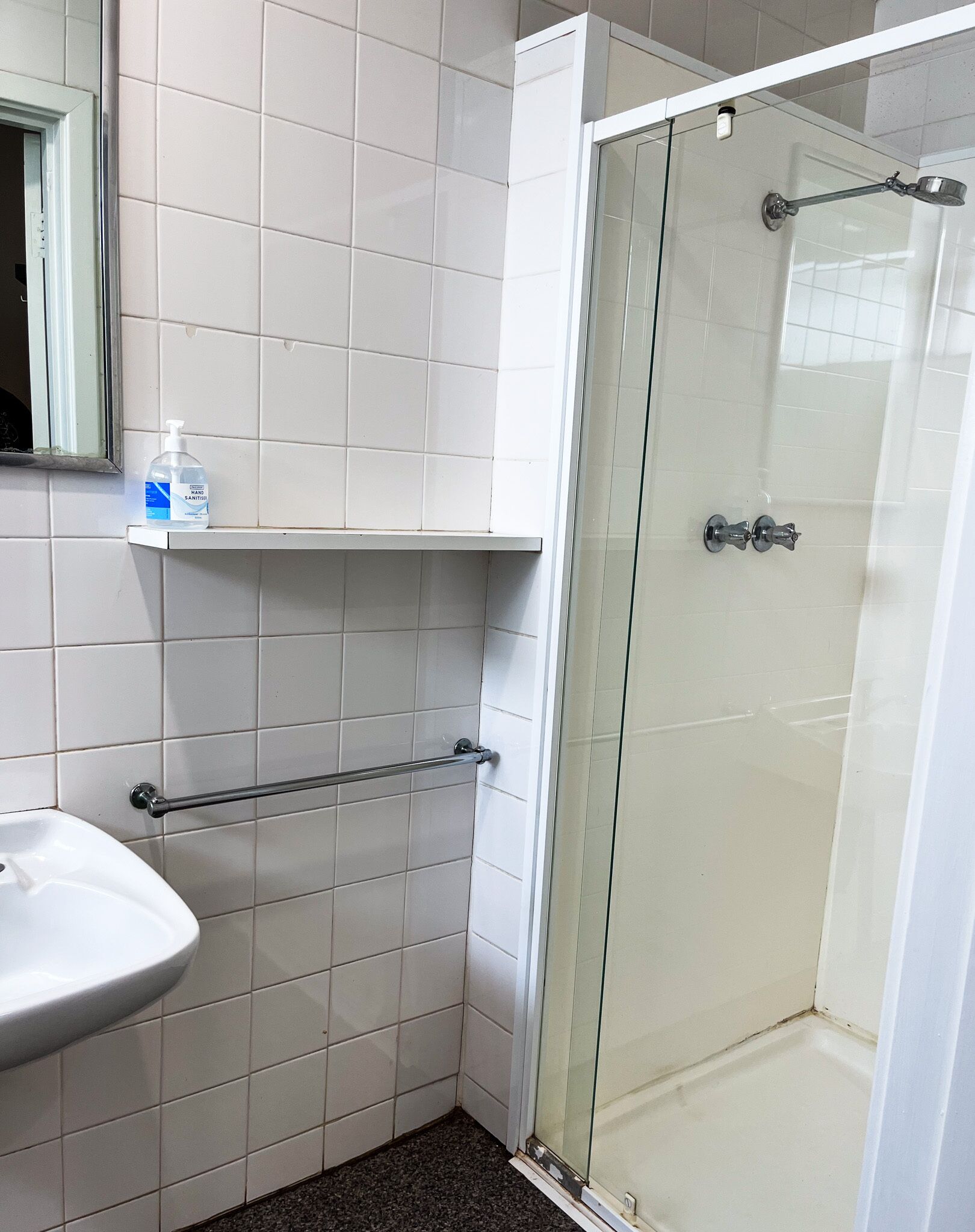 A bathroom with white tiles, a glass-enclosed shower, a white sink, and a shelf with a bottle of hand sanitizer.