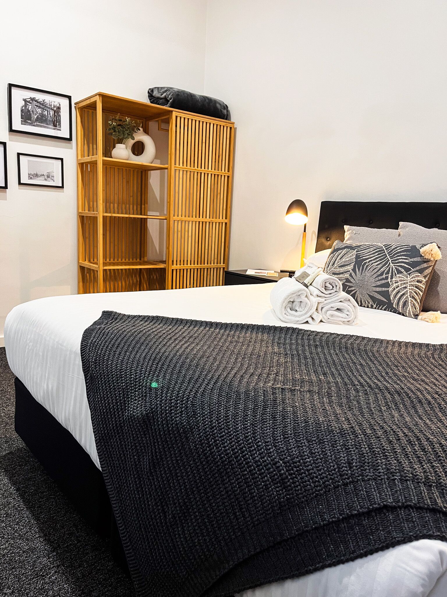A neatly made bed with a black knitted throw, a wooden wardrobe, and a nightstand with a lamp.