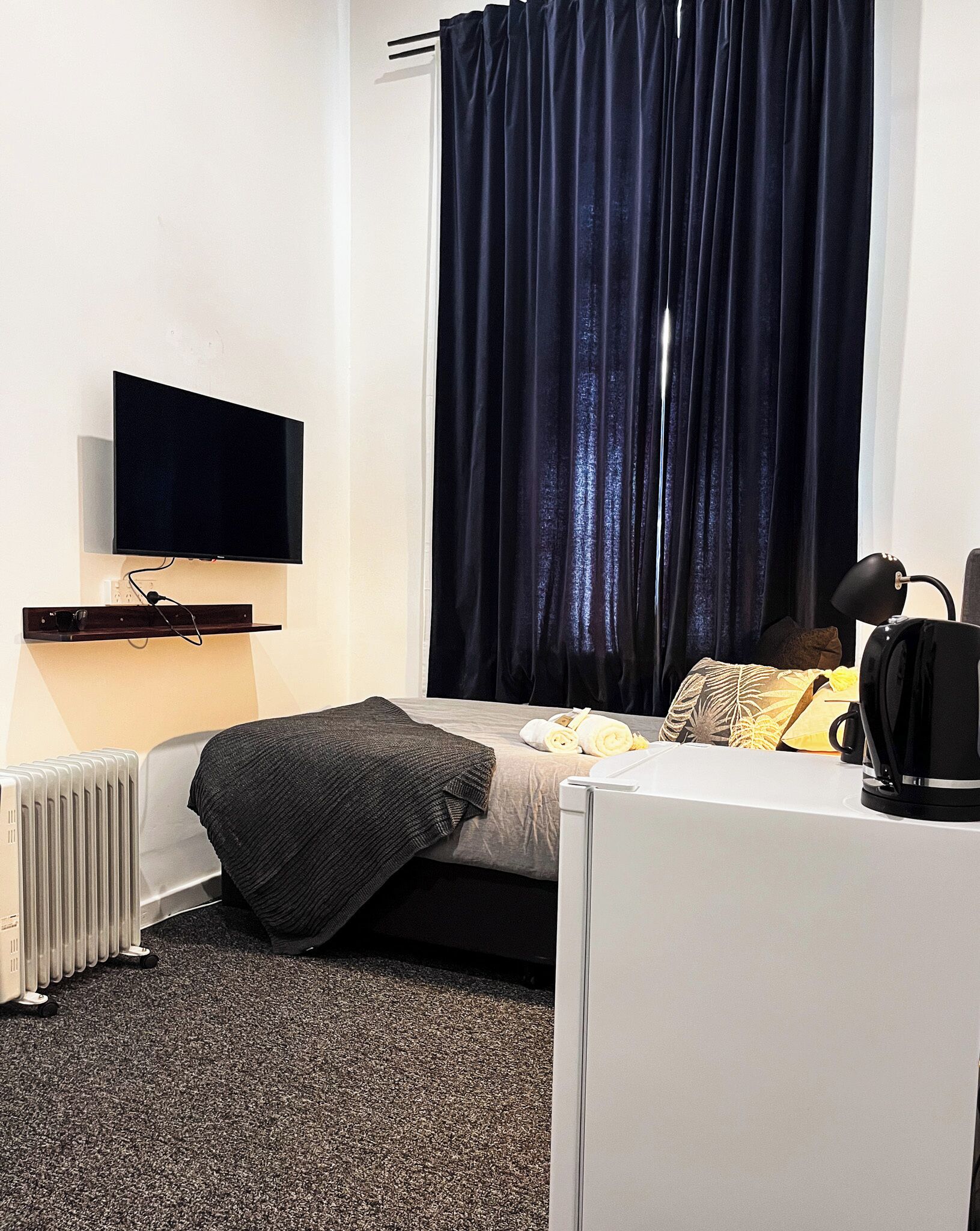 Una camera d'albergo con un letto, una TV a schermo piatto montata a parete, un termosifone, una scrivania con un bollitore e un'ampia finestra con tende scure.