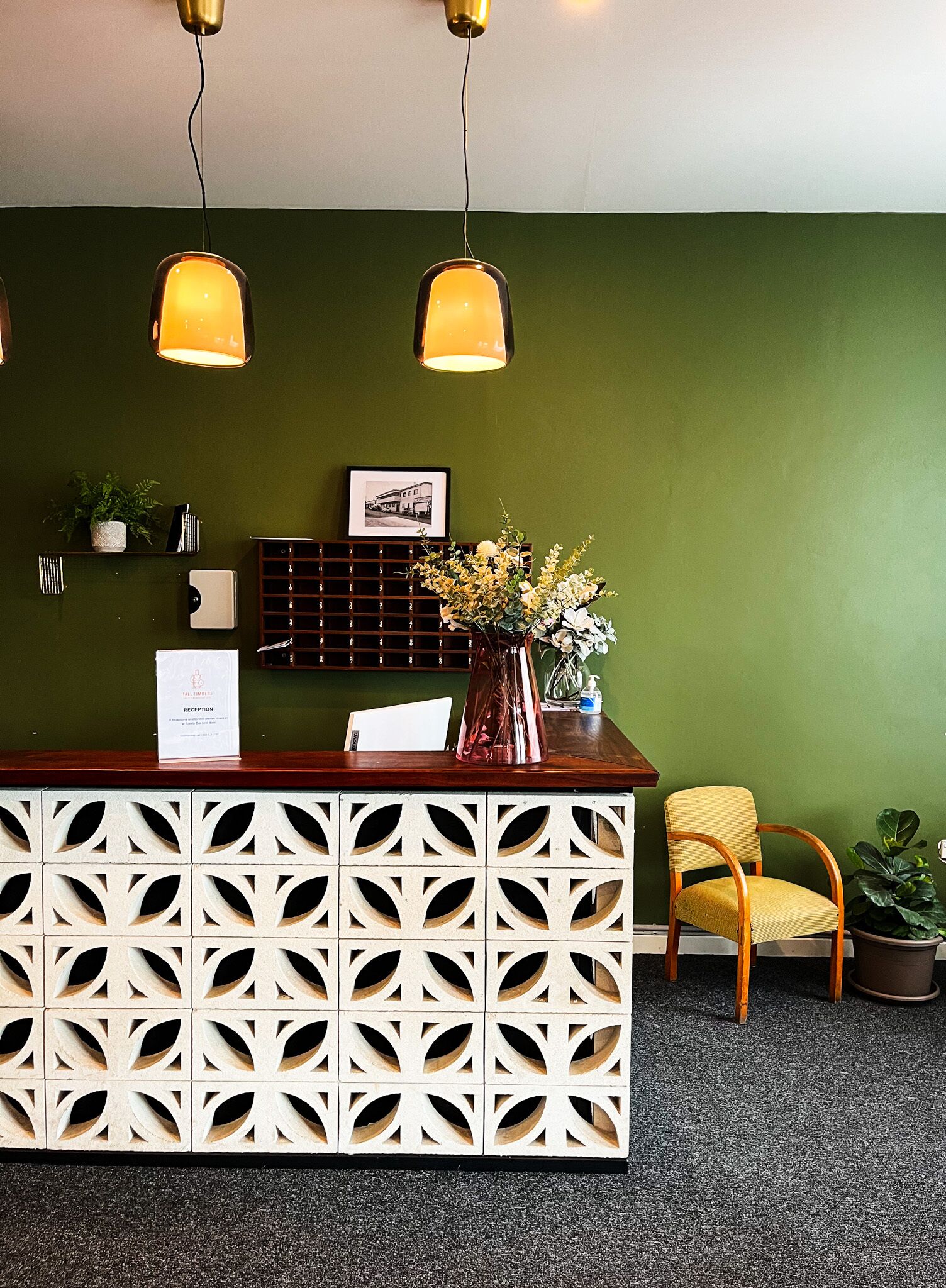 Un mostrador de recepción con una pared verde, un cuadro enmarcado, una planta en maceta y una silla amarilla.