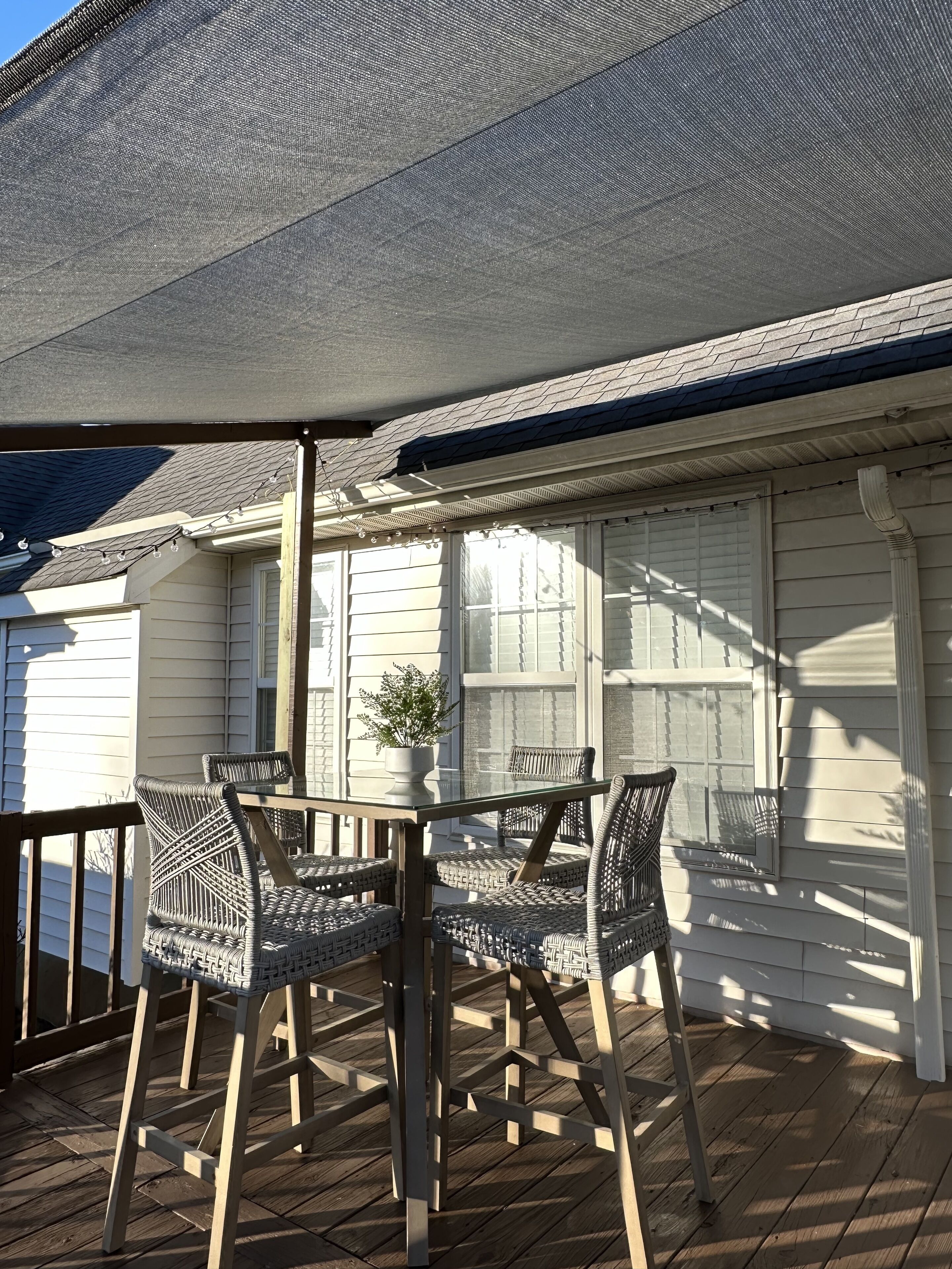 An outdoor patio with wicker furniture and a table, a potted plant, and a house with white siding and a dark roof.