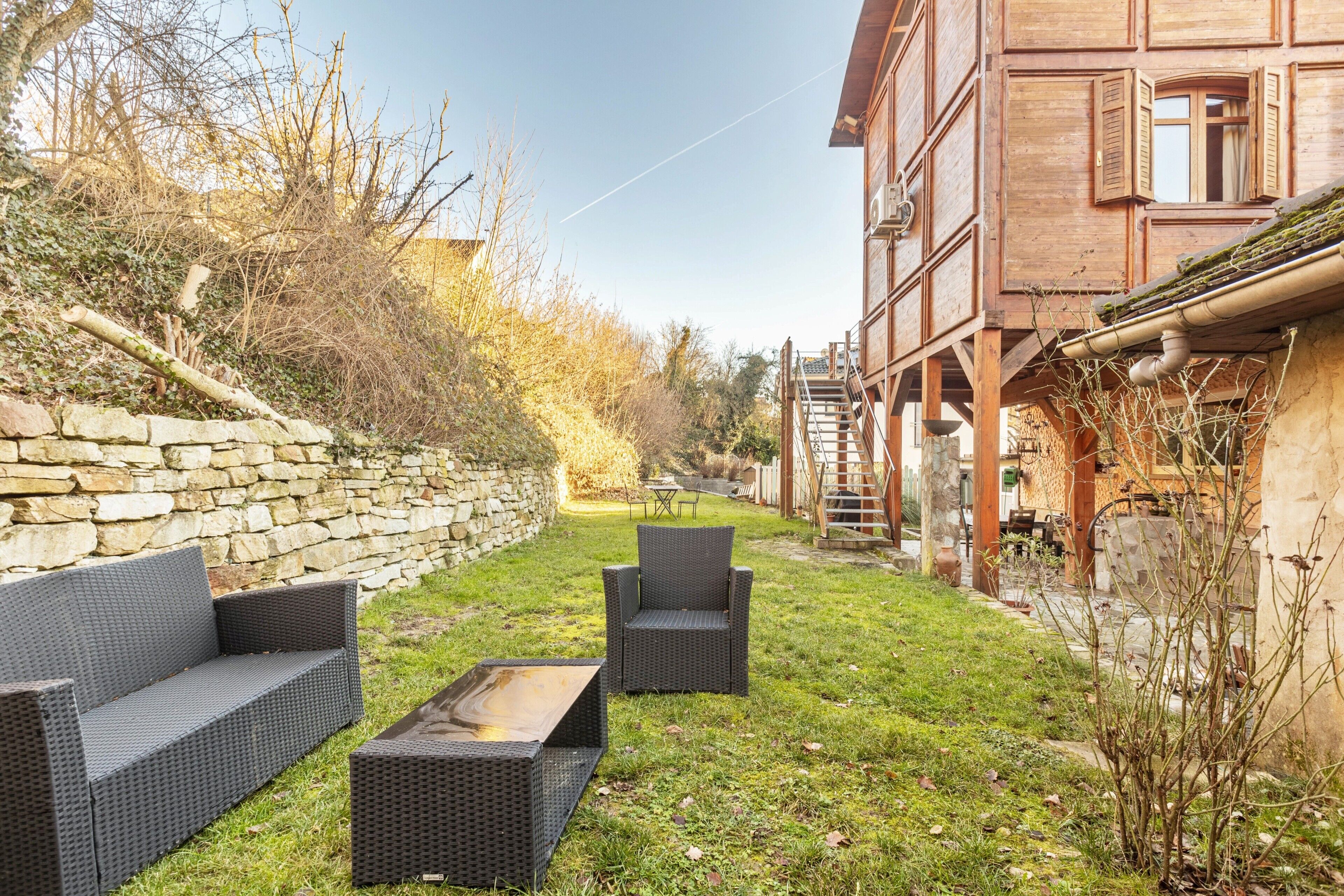 A courtyard with wicker furniture, a stone wall, and a wooden building with windows.