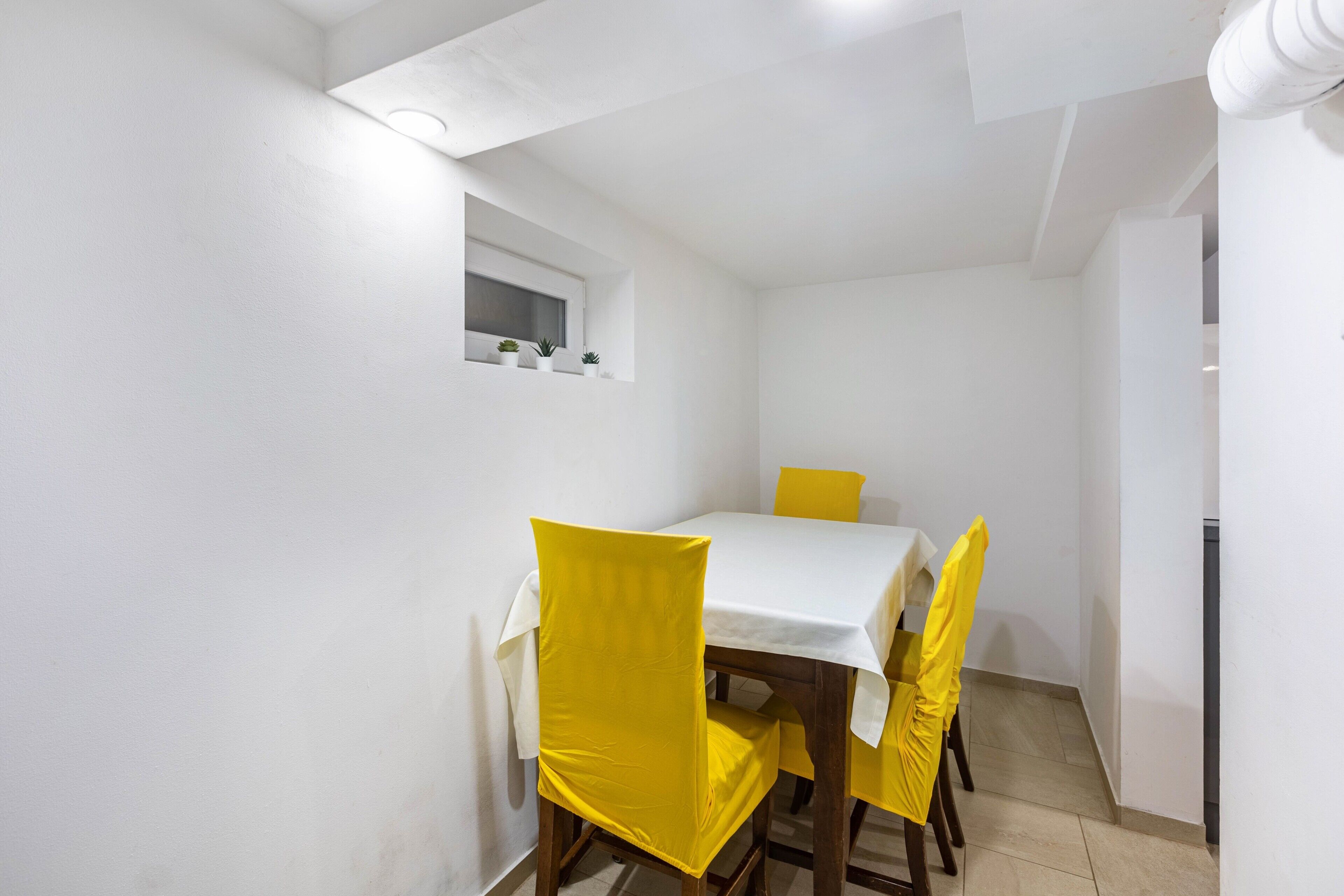 Een gezellig eethoekje met een witte tafel en gele stoelen, een klein raam en een wit plafond.