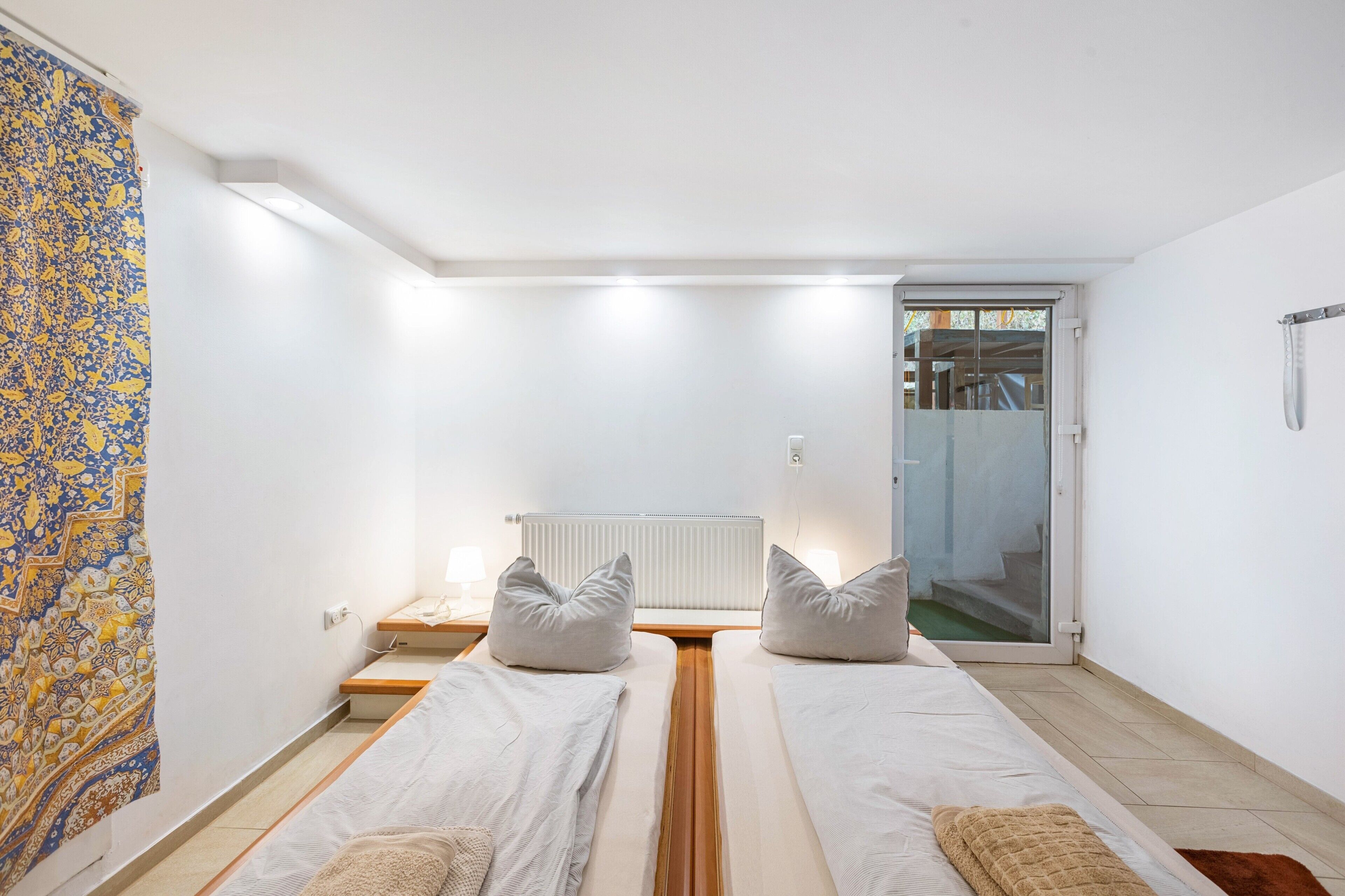 A room with two beds, a radiator, a window, and a door leading to an outdoor area.