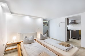 Ein modernes Hotelzimmer mit zwei Einzelbetten, einem kleinen Schreibtisch mit Lampe, einem Heizkörper und einer Küchenzeile im Hintergrund.