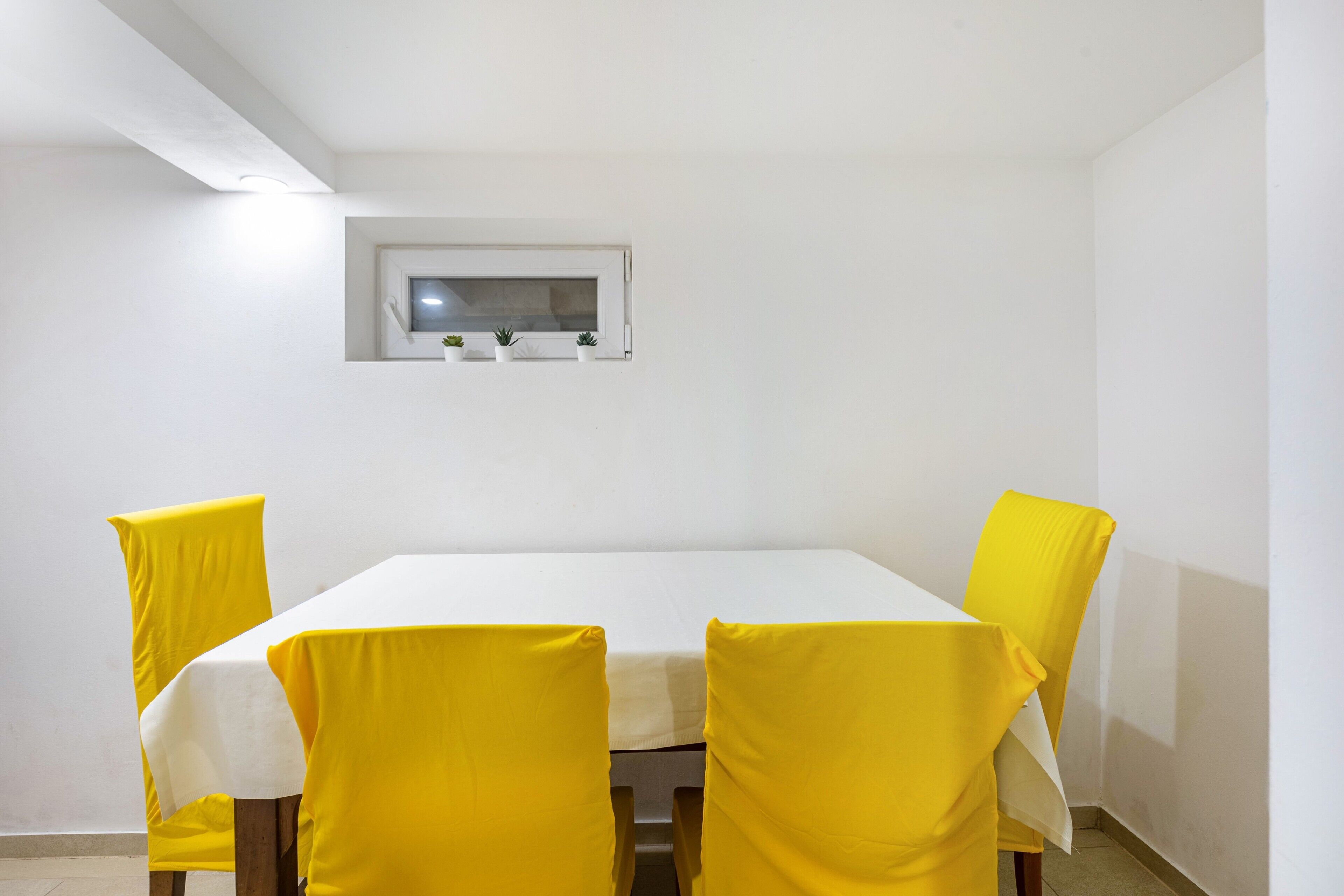 Een eetruimte met een witte tafel, gele stoelen, een klein raam en een lamp.
