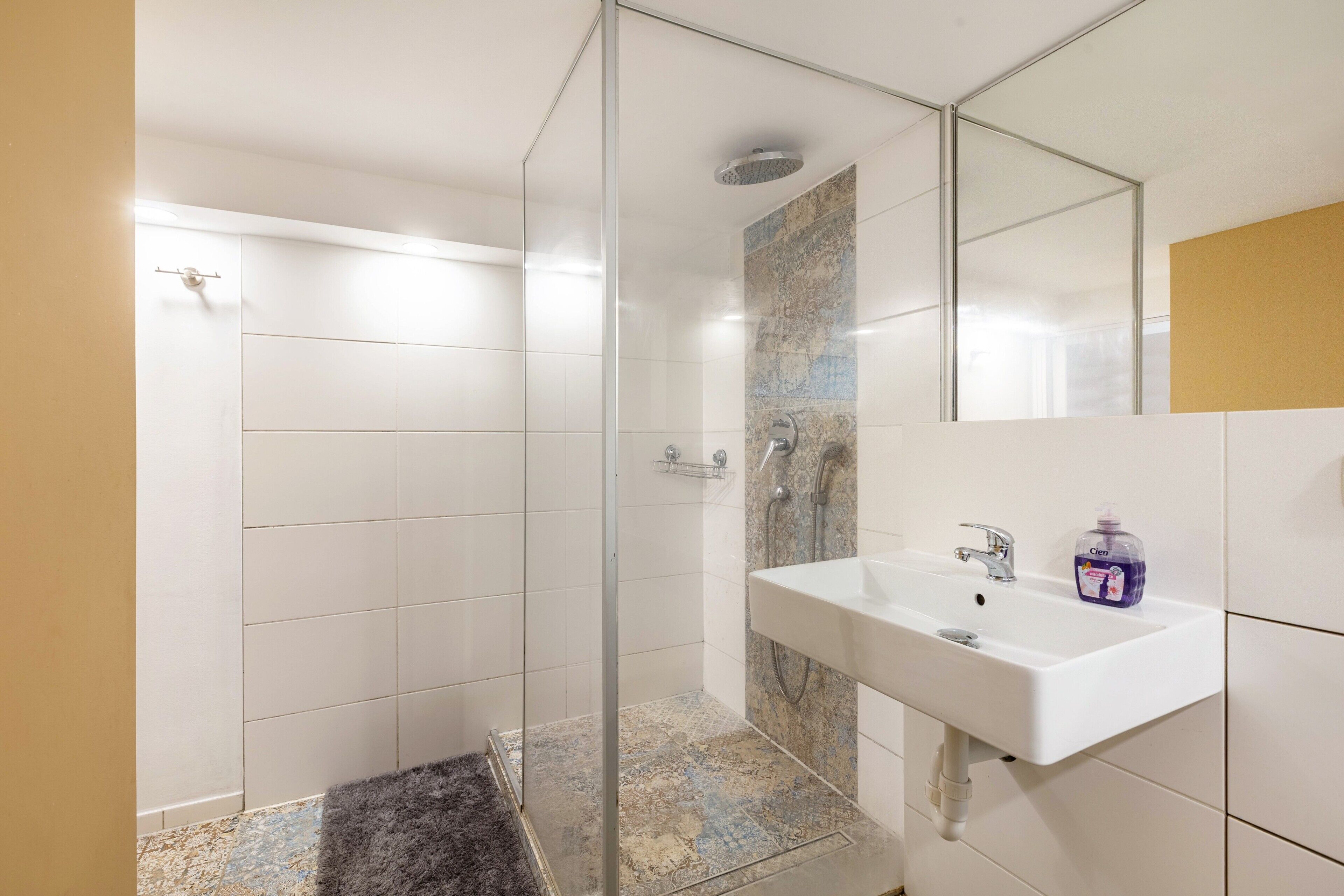 Ein modernes Badezimmer mit einer begehbaren Glasdusche, einem weißen Waschbecken mit Seifenspender und einem Spiegel.