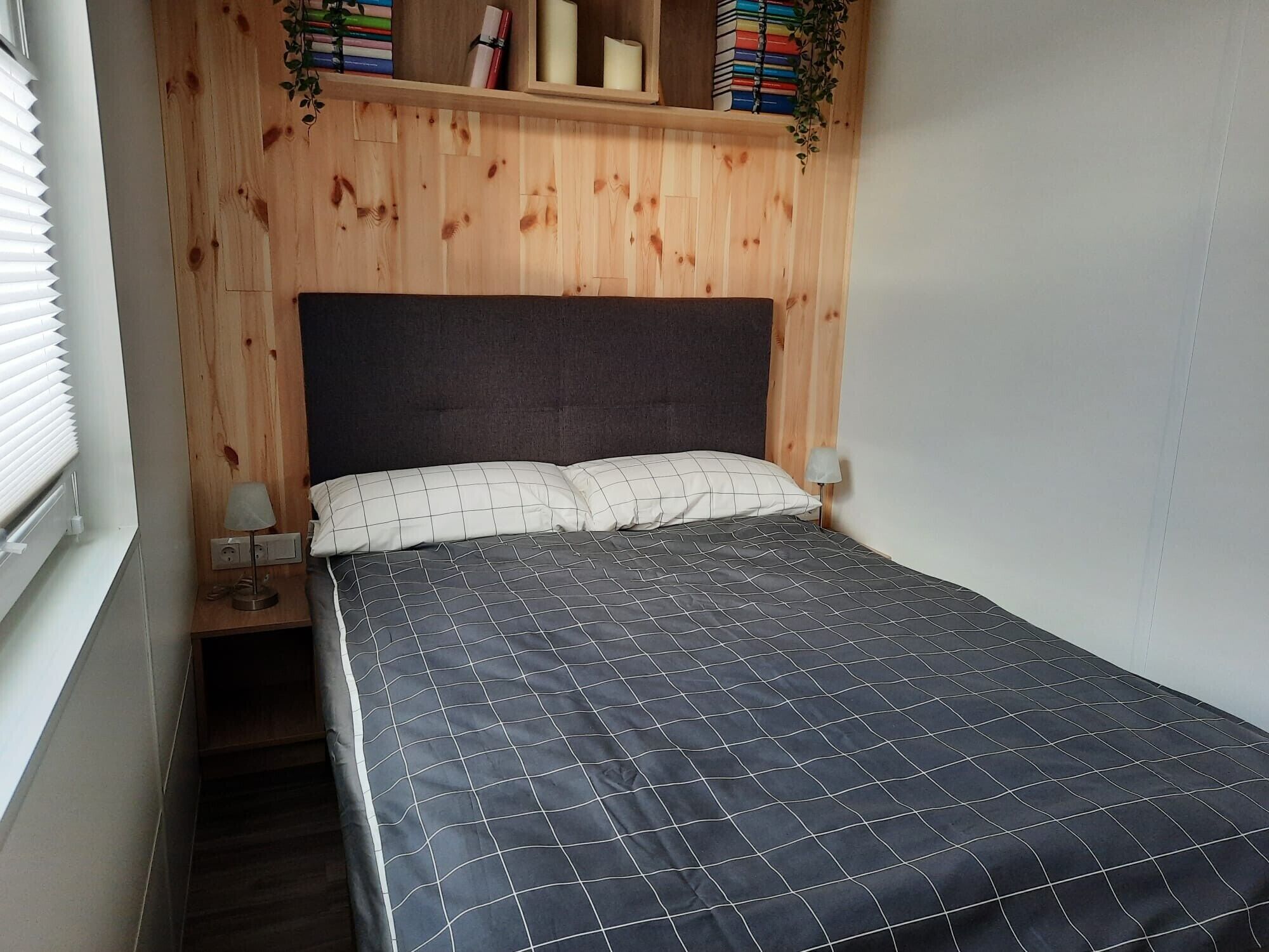 Een slaapkamer met een houten hoofdbord, een bed met een zwart-wit quilt, een nachtkastje met een lamp en een plank met boeken en een plant.
